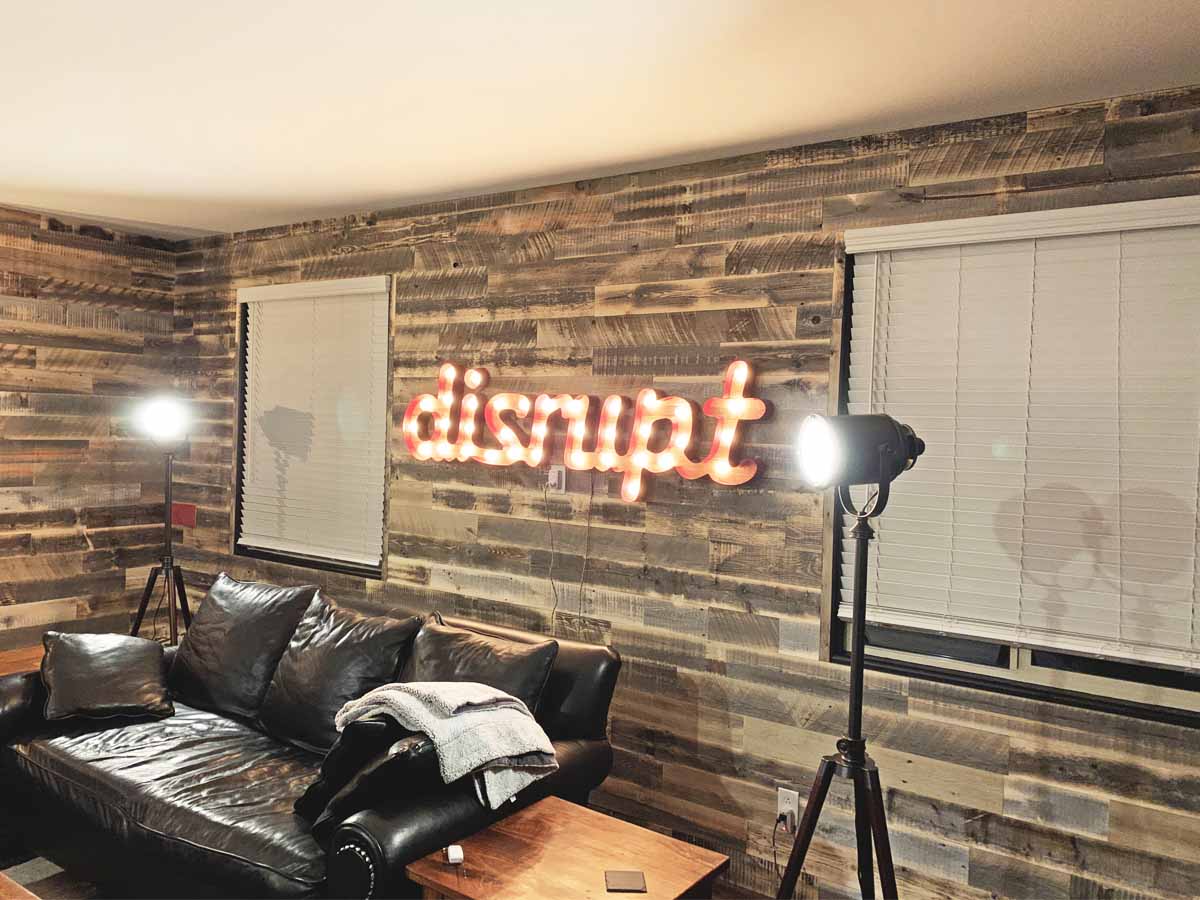 Barn wood wall made from reclaimed snow fence in a rustic living room with a lighted art deco sign that says disrupt.