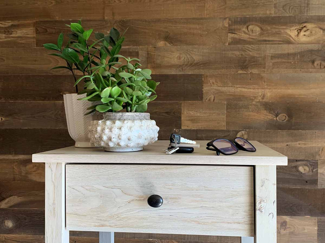 Wooden plank wall in the brown Saratoga finish on a bedroom wall with awhite nightstand with plants, car keys, and glasses.
