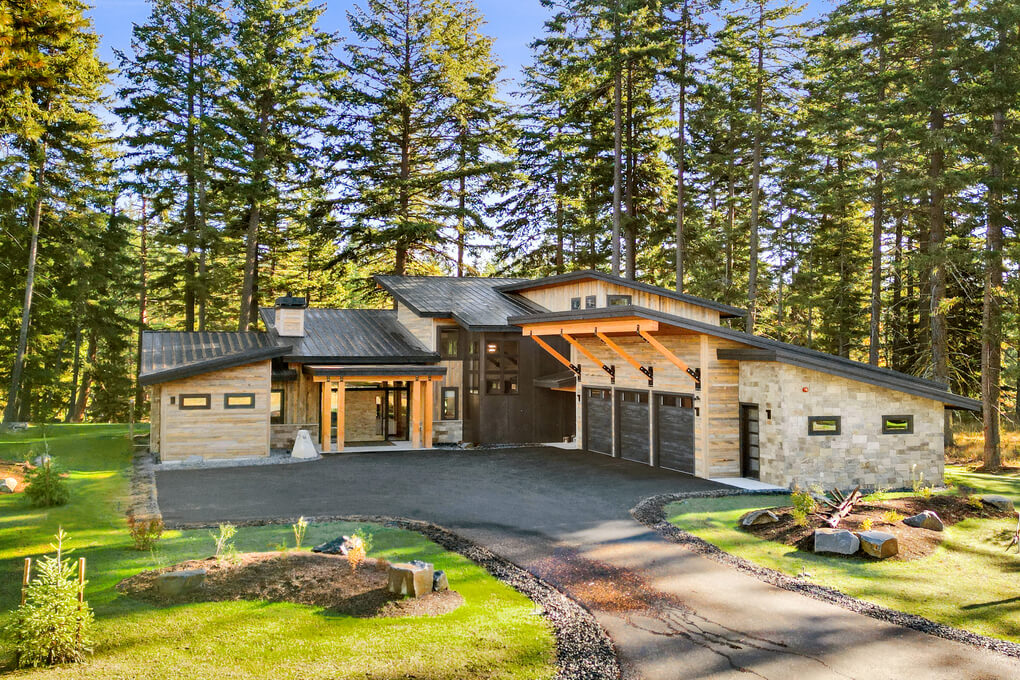 Modern mountain home clad in light colored reclaimed wood siding with a black metal roof set in a stand of pine trees.