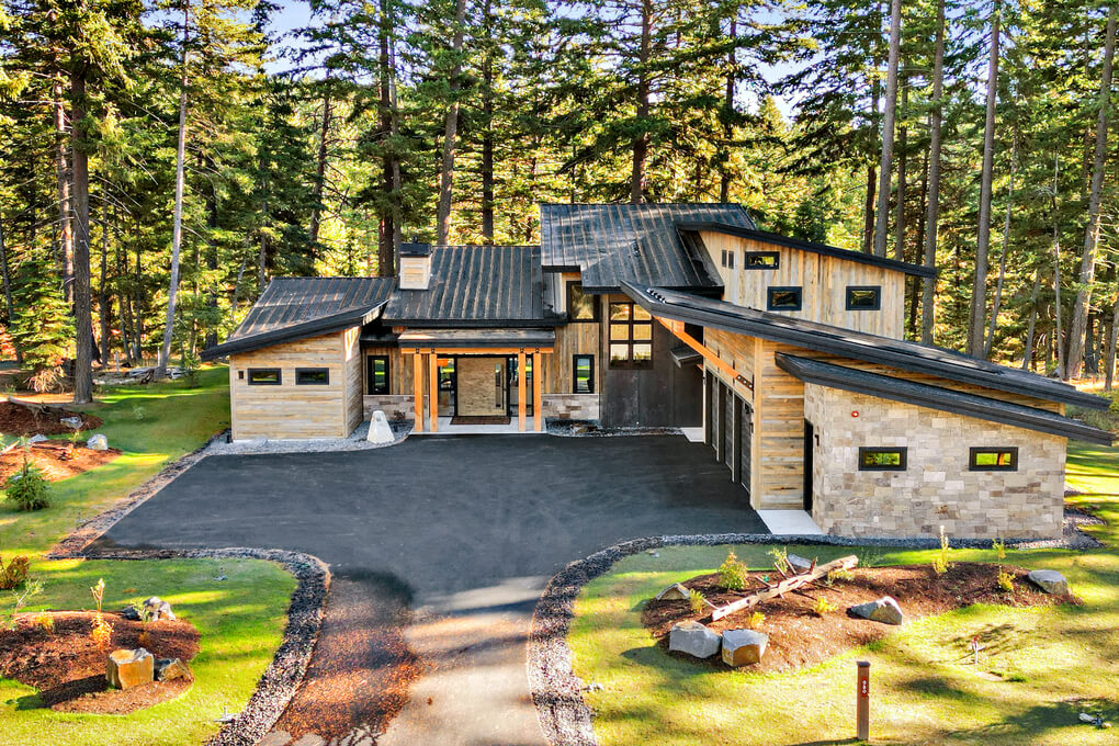 straight on view of a modern mountain home clad in light colored reclaimed wood siding with a black metal roof set in a stand of pine trees.