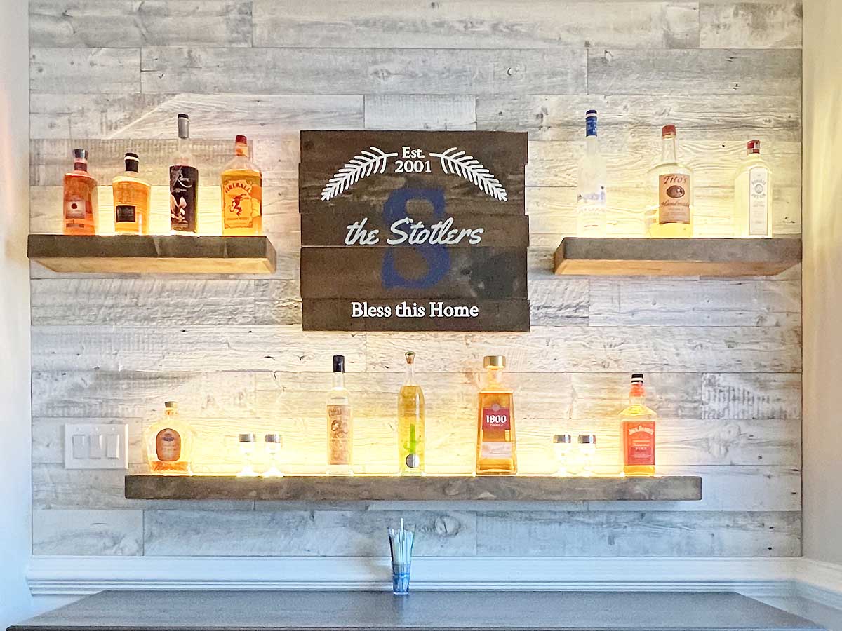 Custom home bar with a whitewashed reclaimed wood background and three floating shelves for bottles.